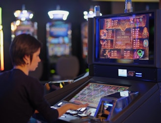 A person is seated at a slot machine console in a casino, focusing intently on the game displayed on the screen. The background shows several other illuminated gaming machines, adding a vibrant and colorful atmosphere to the scene.