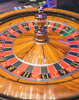 A roulette wheel with alternating red and black numbered pockets on a wooden platform. The wheel contains a small white ball resting in one of the pockets, indicating gameplay. Surrounding the wheel are stacks of colorful poker chips, suggesting a casino environment.