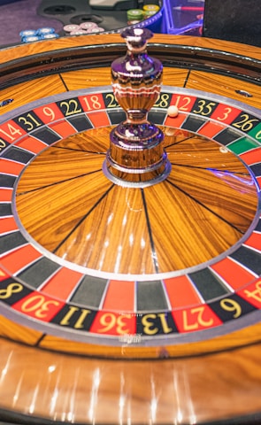 A roulette wheel with alternating red and black numbered pockets on a wooden platform. The wheel contains a small white ball resting in one of the pockets, indicating gameplay. Surrounding the wheel are stacks of colorful poker chips, suggesting a casino environment.